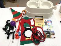 Photo of a large white double dog bowl, assorted dog toys (purple spiky ball, orange bone whistles), red dog collar, red leash, and Christmas-themed dog sweater.