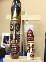 Both hand carved tribal masks displayed upright. Larger mask with prominent wide mouth and geometric facial designs. Smaller mask with red, yellow, white, black painted designs and prominent nose and mouth.