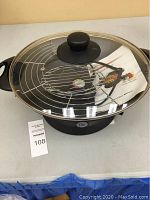 Electric wok with glass lid, metal cooking rack inside, and instruction manual placed on the table. Black base with 'Elite' brand visible.