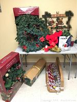 Full view of table with 4 pine wreaths and garlands, artificial pine tree in box, candy cane string lights in box, and small Noel decoration