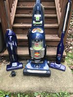 Three vacuums shown together outside: two upright Bissell PowerTrak Revolution models and one Oreck handheld model with charging base.