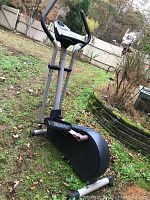 Outdoor photo showing elliptical exercise machine on grass, including frame, pedals, handlebars, and console display.