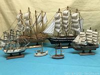 Wide angle photo showing all six wooden sailing ship models arranged on a blue cloth background, showcasing three larger and three smaller boats with detailed sails and rigging.
