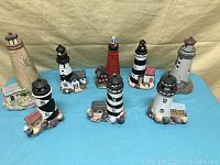 Eight lighthouse sculptures including a tan brick lighthouse music box and seven other painted lighthouse figures on a blue cloth background.