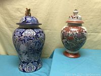 Photo showing both vases side by side, blue one with gold edging and lid with gold finial on left, white and red floral urn with lid on right.