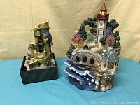 Two decorative fountains shown together on a blue surface with a plain tan background: the bluebird bamboo fountain and the lighthouse village fountain.