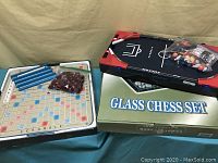Scrabble game board with tile racks, letter tiles bag, multi-sports game board, and box labeled Glass Chess Set