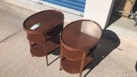 Pair of wooden oval end tables with glass tops, two lower wooden shelves, medium brown finish, outdoor daytime photo.