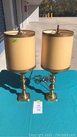 Pair of brass lamps with beige drum shades on a blue cloth surface displayed outdoors