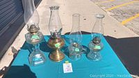 Four glass hurricane lamps arranged on a blue table cloth outside, showing varied bases and brass burner assemblies.