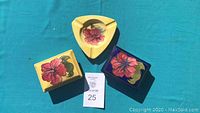 Top view of three Moorcroft pottery pieces with hand-painted red hibiscus flowers on yellow and blue backgrounds, arranged on turquoise surface.