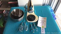 Sunbeam Mixmaster stand mixer head with 4 attachments and 2 nested mixing bowls on a blue surface, with instruction manual visible beside the mixer and mixer base.
