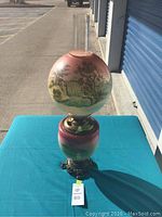 Full view of antique Gone With The Wind lamp on blue cloth showing hand-painted waterfall scene globe and matching font with glass chimney and decorative metal base.