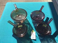 Four transparent amber glass pots with handles and clear lids arranged on blue tablecloth in sunlight.