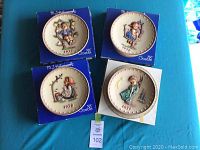 Four M.J. Hummel decorative plates arranged on blue boxes showing the front designs and years 1971, 1976, 1977, and 1978.