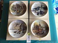 Four decorative plates arranged in a square, each illustrating a different rural or small town scene with people, buildings, and natural elements in various seasons. Plates are 10 inches in diameter.