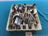 Overhead photo of assorted stainless steel flatware including knives, forks, and spoons in a white plastic flatware tray on a blue surface.