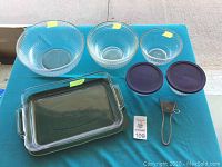 Overview photo showing three clear ribbed glass Pyrex mixing bowls, two purple plastic lids, rectangular Anchor Hocking baking dish with lid, and metal utensil on a teal table.