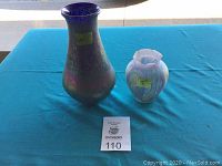 Photo showing two handblown vases on blue cloth, one larger iridescent purple-blue vase and smaller white vase with pattern