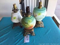 Three antique electrified oil lamps on blue table showing painted glass bodies and ornate metal bases