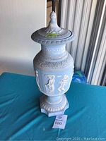 Front view of a 20-inch tall light blue ceramic lidded urn with white classical relief figures and ornamental design, standing on a pedestal base.
