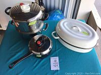 Full set of pots and pans on blue cloth including a large stainless steel pot with glass lid, a black saucepan with lid and a white enamel roasting pan with lid.