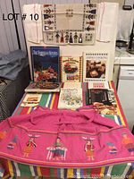 Photo of 8 international cookbooks spread beside ethnic kitchen textiles including a pink apron with folk embroidery, a patterned tablecloth, tea towel, and hand towels hung above
