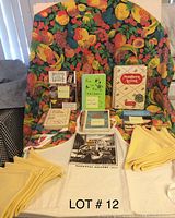 Cookbooks arranged on and around a colorful fruit-themed tablecloth seated on a chair, surrounded by yellow napkins and a yellow apron all visible in the image.