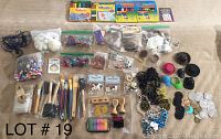Top-down view of assorted craft supplies organized on a surface. Includes bags of beads, paint brushes, stamps with wooden handles, ink pads, pom poms, googly eyes, feathers, colorful markers, and various decorative trims.