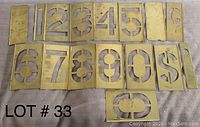 Photo showing brass stencils laid out on a surface. The numbers 1 through 9, 0, a dollar sign, and two blank brass plates are visible. Each stencil measures approximately 6 inches tall.