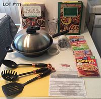Full set showing Turbo Cooker Plus with lid, three utensils, chopping mat, manual chopper, measuring cup, two hardcover cookbooks, and three recipe booklets on table