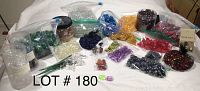Photo shows a collection of assorted colorful floral stones and glass beads spread on a white surface. Various colors including green, clear, yellow, purple, red, blue, black, and mixed colors visible. Multiple containers and zip bags hold the stones. Floor and background partially visible.