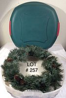 Photo showing artificial evergreen Christmas wreath with pinecones placed in front of a matching green round wreath storage container with red side handles.