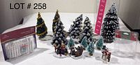 Photo showing 7 Christmas trees, 3 figurines, 3 tree-shaped candles in box, and a white snow fence.