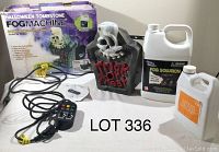 Halloween-themed fog machine shaped like a tombstone with skull, alongside two plastic jugs labeled as fog juice and a remote control with attached power cord, and original box in background.
