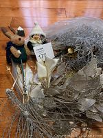 Photo of assorted Christmas ornaments and plush figures including a bunny and penguin, various metallic floral decorations, ribbons, and silver tinsel garland arranged on a wooden floor.