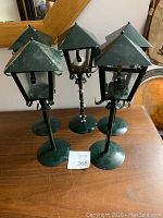 Five green metal Tamp post style tea light holders with glass panes and curled metal accents, arranged on a wooden surface.