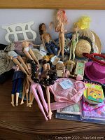 Overview of multiple fashion dolls, miniature furniture pieces, assorted hats, clothing items, and small children's books on a wooden surface.