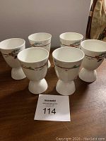 Six white Royal Doulton bone china egg cups displayed on a table, each decorated with a subtle pink and green floral vine band around the rim.