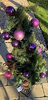Full view of the green artificial pine topiary with purple baubles and pine cones set on a brown base outdoors.