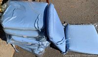 Stack of four light blue outdoor chair cushions wrapped in plastic, including seat and back cushions, showing folds and wear.