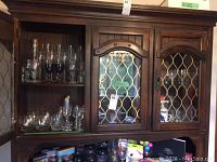 Wooden cabinet with double doors holding multiple rows of clear and tinted glass stemware including various types like beer and wine glasses.