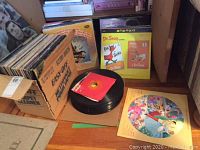 Overall view of cardboard box filled with LP sleeves, pile of 45 rpm singles, and children’s albums