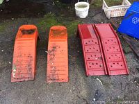 Two sets of auto ramps: one set orange plastic ramps, and one set red metal ramps, placed side by side on ground with dirt and moss
