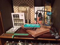 Photo of overall lot including boxed cutlery sets, carving board, metal skewers, knife and some kitchen accessories all on wooden shelf.