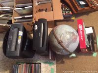 Photo showing two black paper shredders, a globe, Dymo tape label maker in red box, and various bundled papers and boxes in background