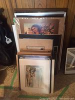 Stack of various framed and matted artwork leaning against a wall, includes a visible female portrait and landscape prints