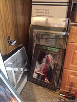 Photo showing several framed prints stacked and leaning in a corner, including a partially visible large black-and-white art print and a colorful Native American portrait.