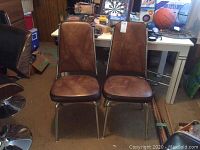 Two brown retro kitchen chairs with padded seats and backs, metal chrome legs, placed on brown carpet in a room with other items.