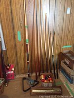 Photo showing multiple wooden pool cues leaning against a wood-paneled wall, one detachable into 4 sections, with a box of billiard balls on the floor.